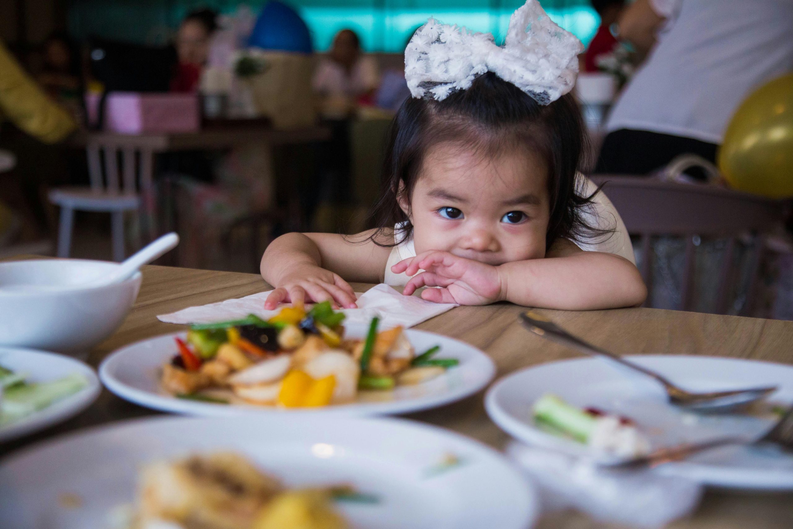 Adorable toddler with a large bow waiting at a table filled with colorful food at a party.