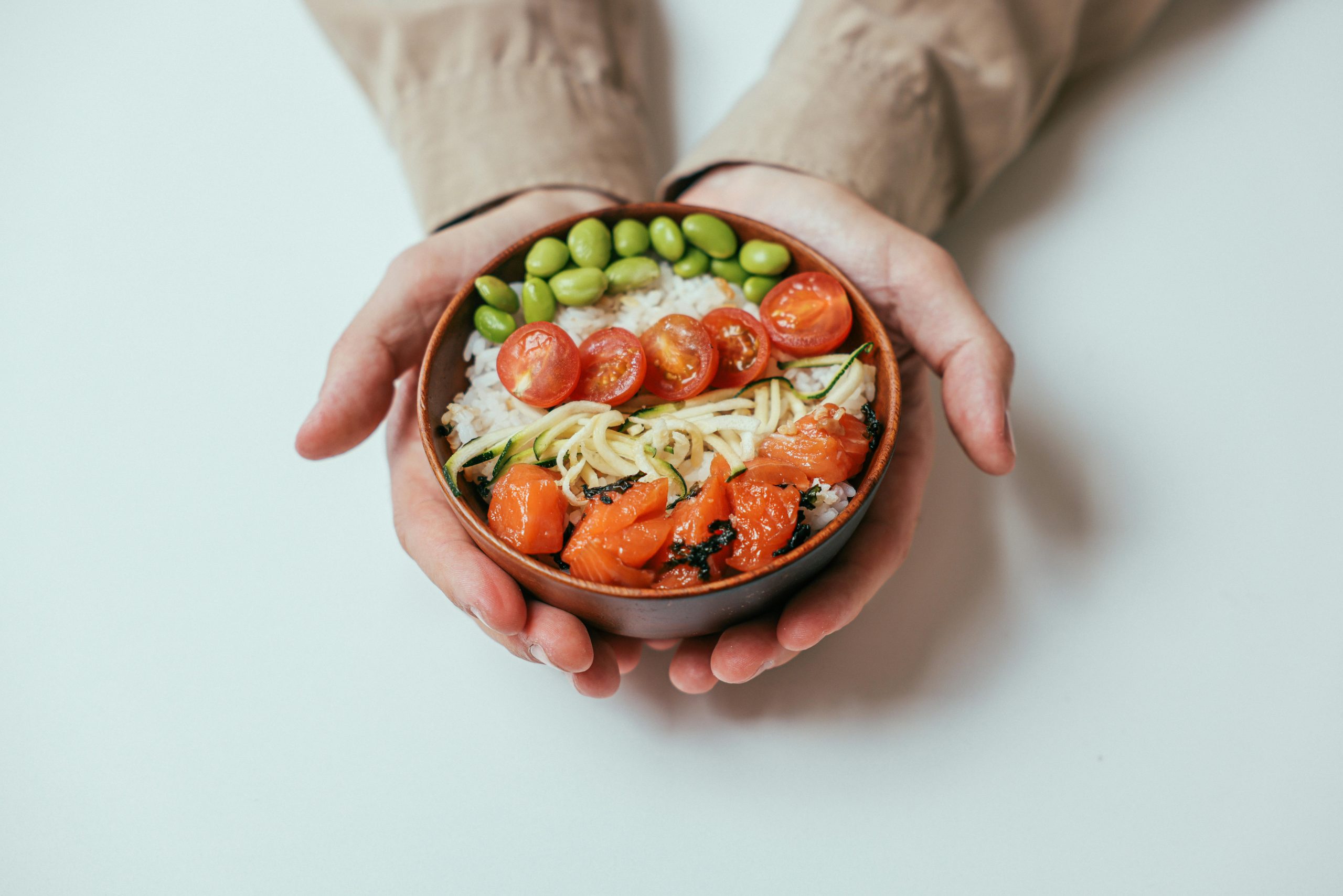Close-up of hands holding a salmon poke bowl with cherry tomatoes, edamame, and rice.