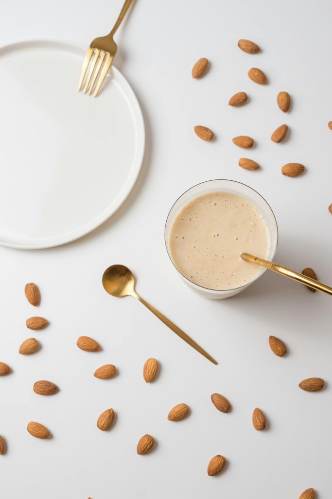 Stylish flat lay of almond milk with scattered almonds and golden cutlery on white surface.